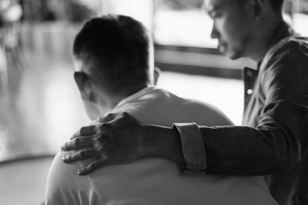 Rear view of son and elderly father sitting together at home. Son caring for his father, putting hand on his shoulder, comforting and consoling him. Family love, bonding, care and confidence