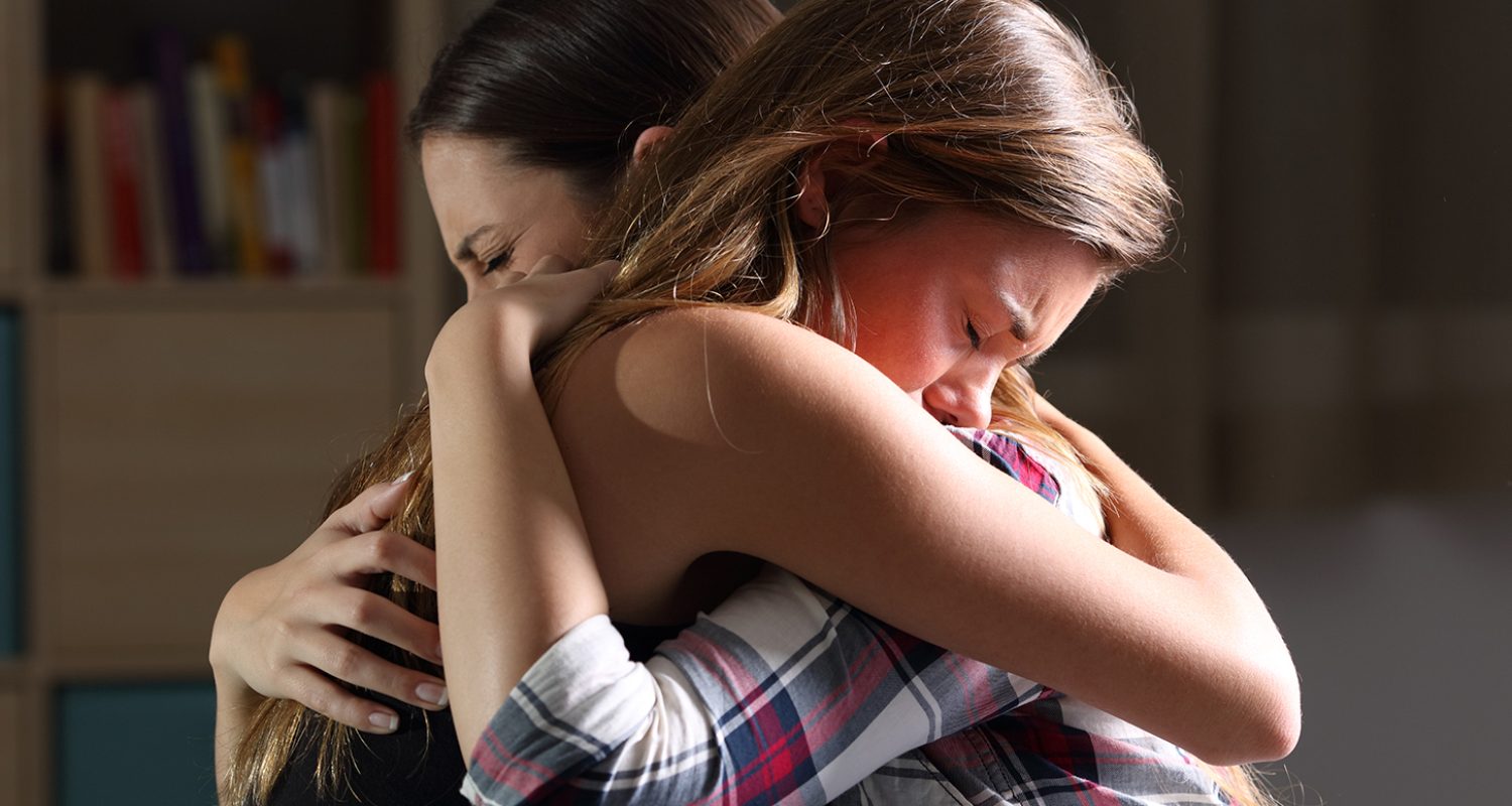 Side view of two sad good friends embracing in a bedroom in a house interior with a dark light in the background