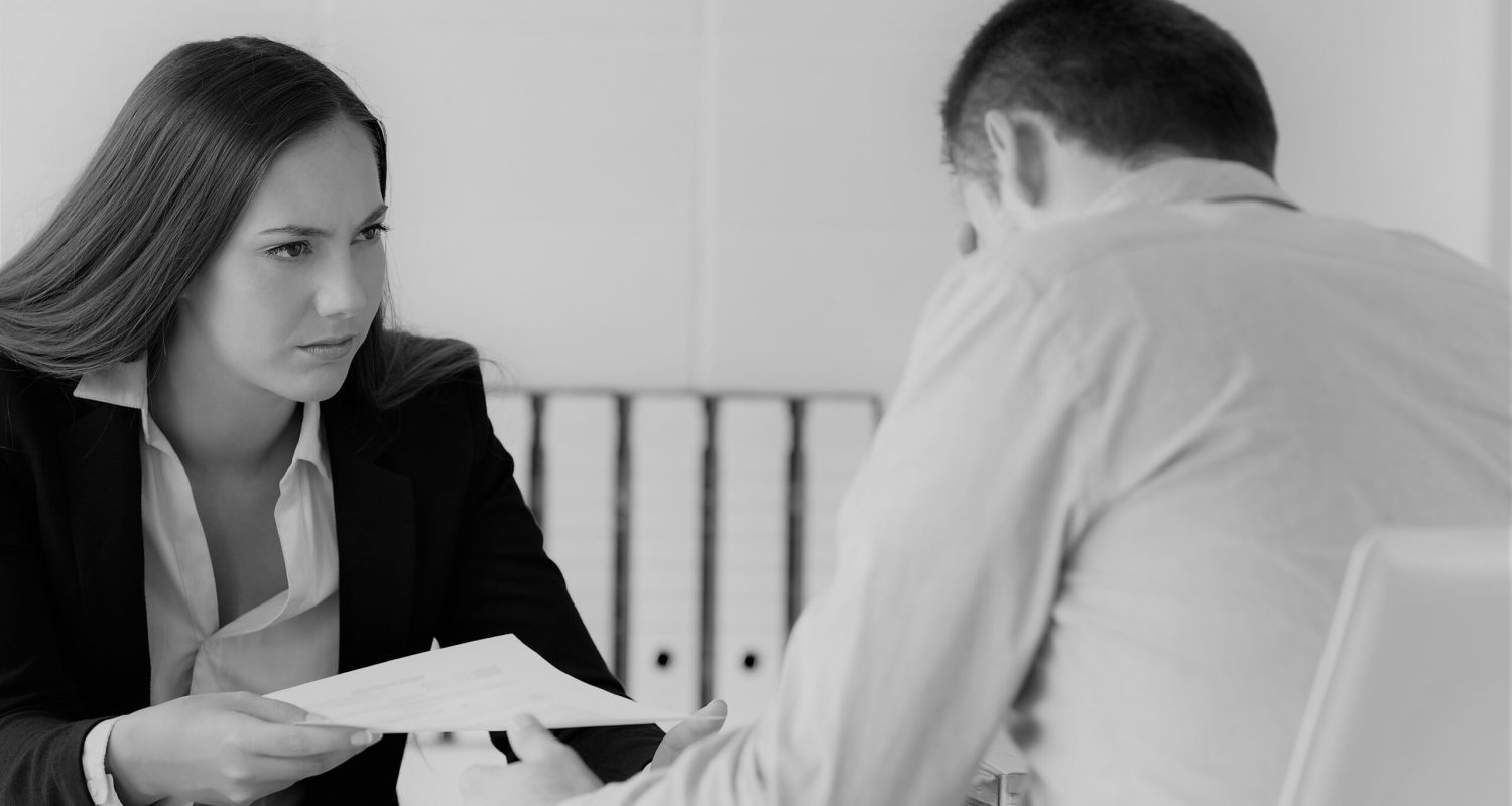 Angry boss giving a document to a sad employee sitting in a desk at office