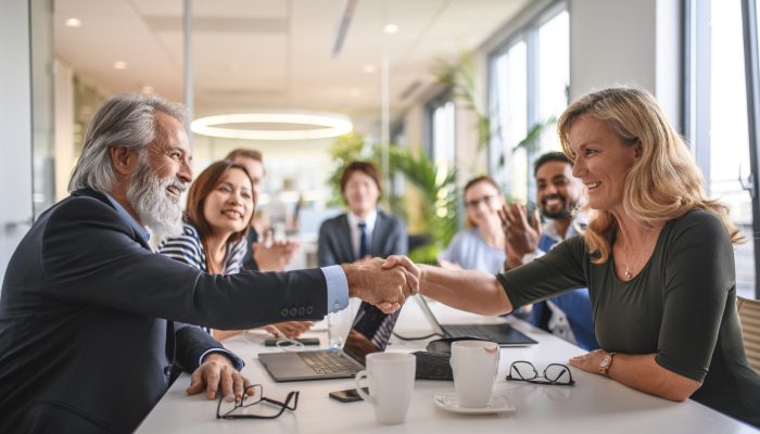 Close-up of smiling male and female business associates celebrating success with a hand shake as colleagues look on.