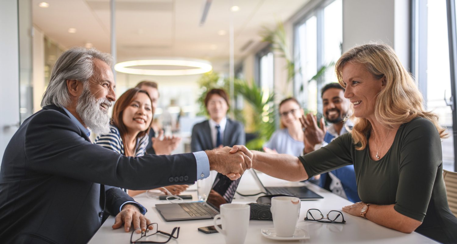 Close-up of smiling male and female business associates celebrating success with a hand shake as colleagues look on.