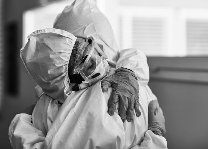 Male and female medical coworkers wearing protective suits, eyewear, and gloves hugging in support and recognition of hard work during COVID-19 pandemic.