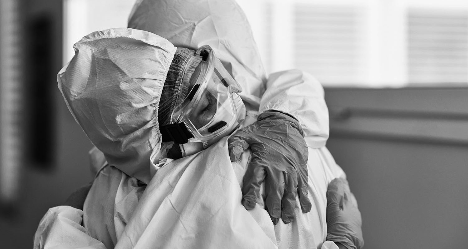 Male and female medical coworkers wearing protective suits, eyewear, and gloves hugging in support and recognition of hard work during COVID-19 pandemic.