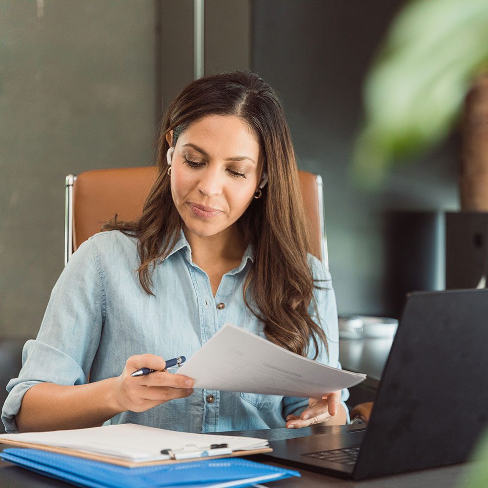 A mid adult businesswoman reviews documents while working in her office. An open laptop as well as a desktop pc are on her desk.