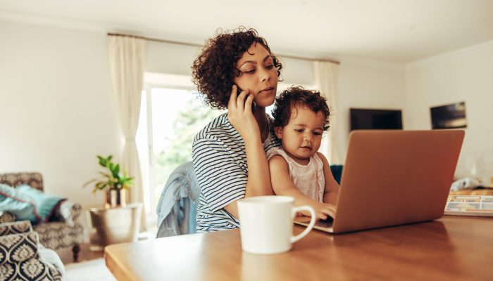 Woman freelancer using cellphone and laptop with her baby sitting on her lap at home. Working mother at home with baby.