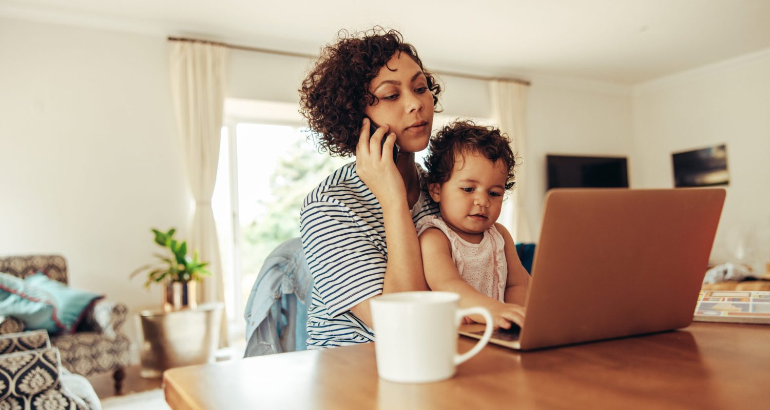 Woman freelancer using cellphone and laptop with her baby sitting on her lap at home. Working mother at home with baby.