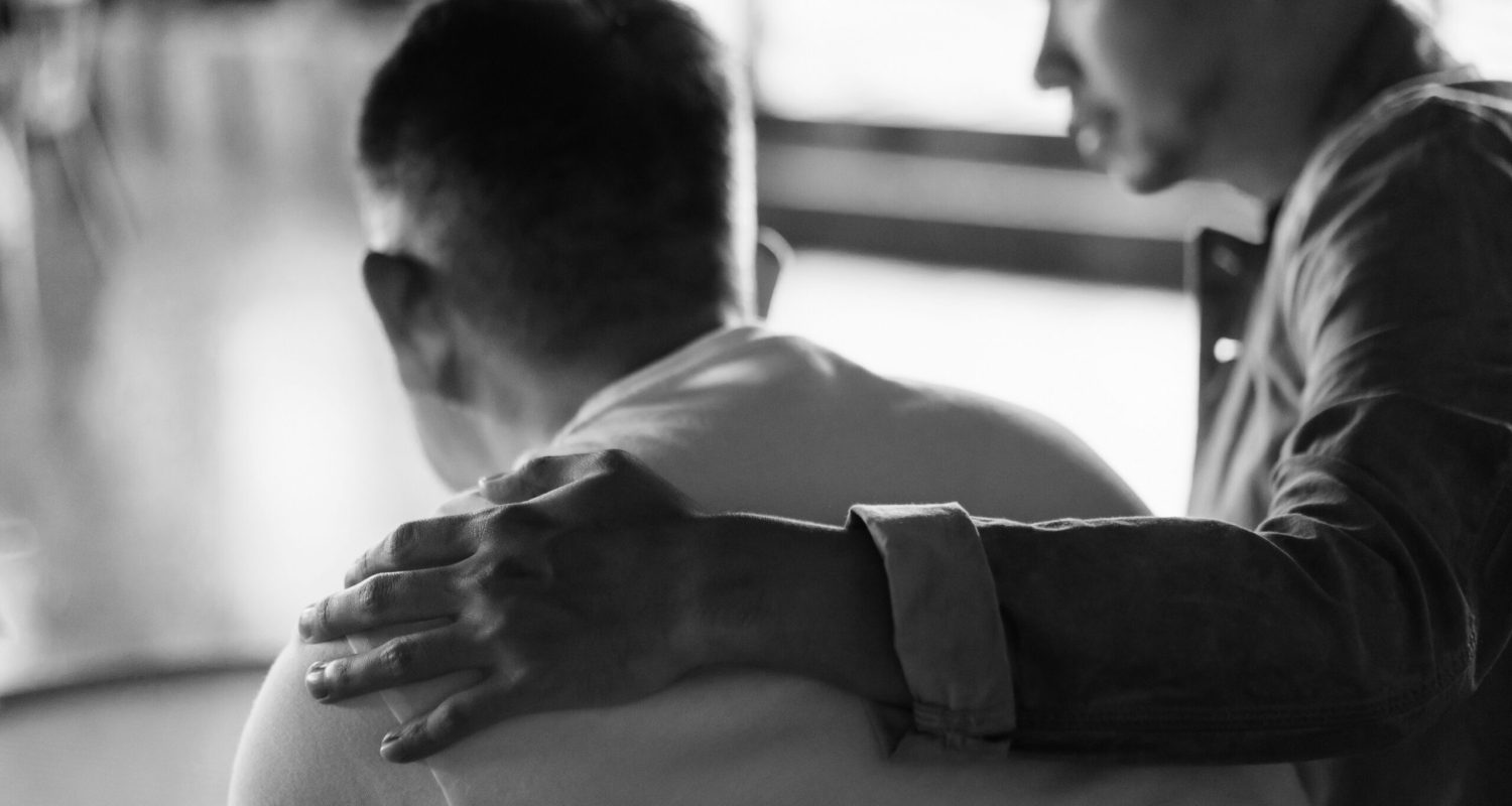 Rear view of son and elderly father sitting together at home. Son caring for his father, putting hand on his shoulder, comforting and consoling him. Family love, bonding, care and confidence