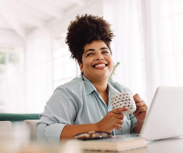 Wide angle shot of woman sitting on table with coffee mug and laptop. Woman taking break while working from home.