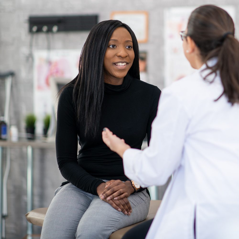 A middle aged woman of African decent, sits up on an exam table as she talks with her doctor about her health concerns.  She is dressed casually and her female doctor is seated across from her in a white lab coat as the two carry on a discussion.