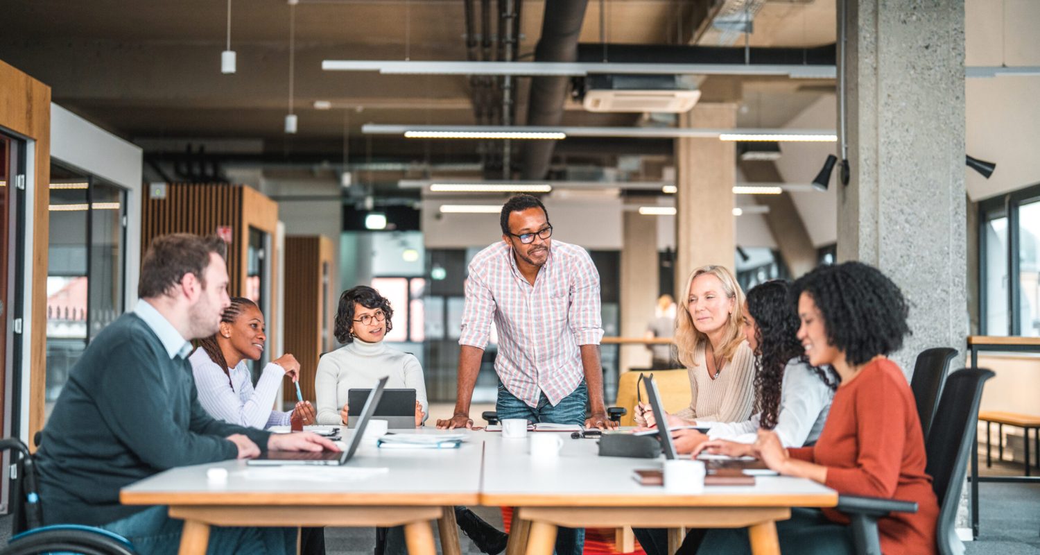 Bright open concept office. There is a work meeting being held where a group of diverse people, including a man on a wheelchair are working together.