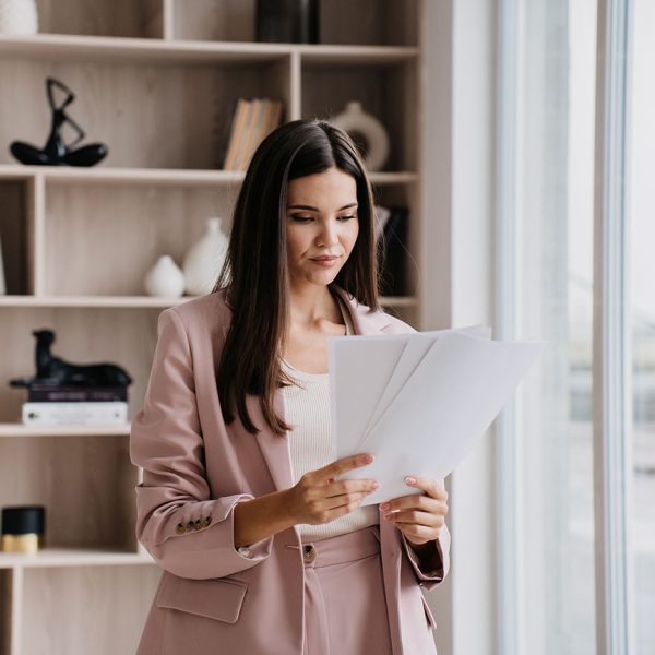 Brunette Asian businesswoman in suit holds sheet of papers looks at documents stands at window against bookshelves. Successful lawyer explores issue at office. Financial bill.