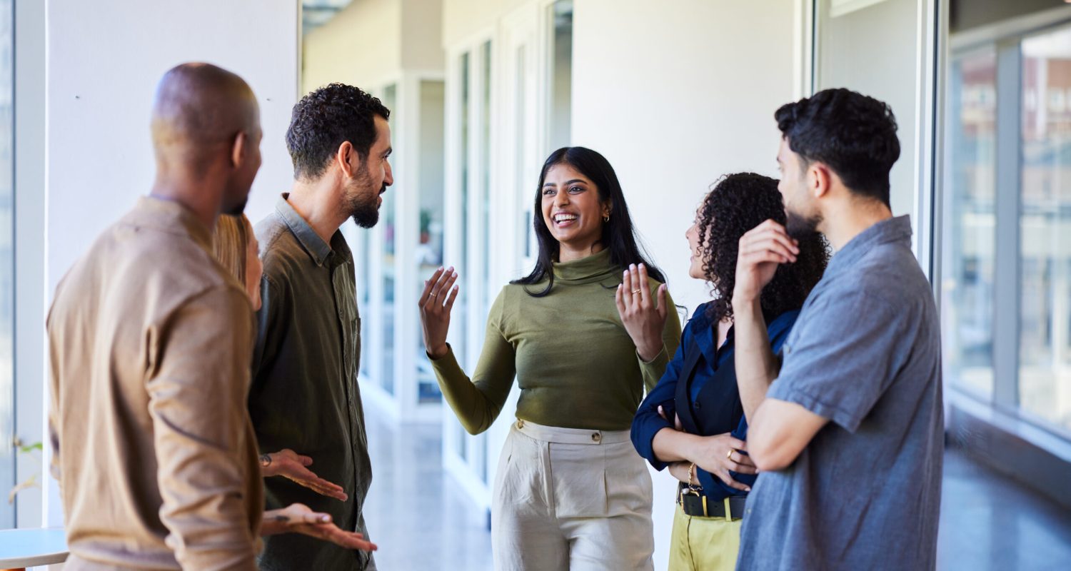 Young businesswoman talking and laughing with a group of diverse coworkers while standing together in the corridor of an office