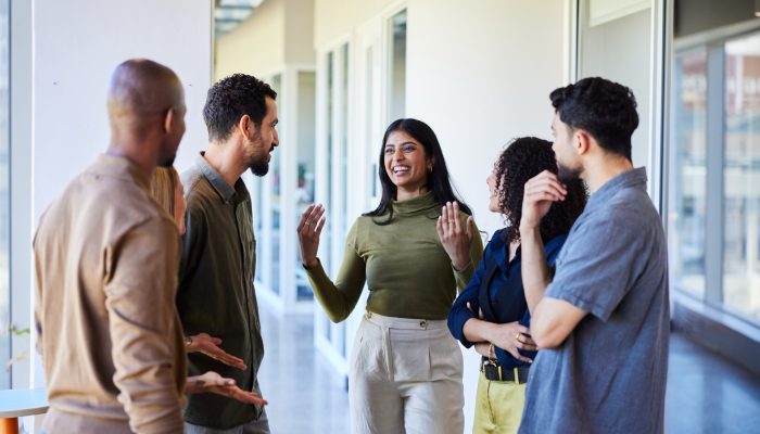 Young businesswoman talking and laughing with a group of diverse coworkers while standing together in the corridor of an office