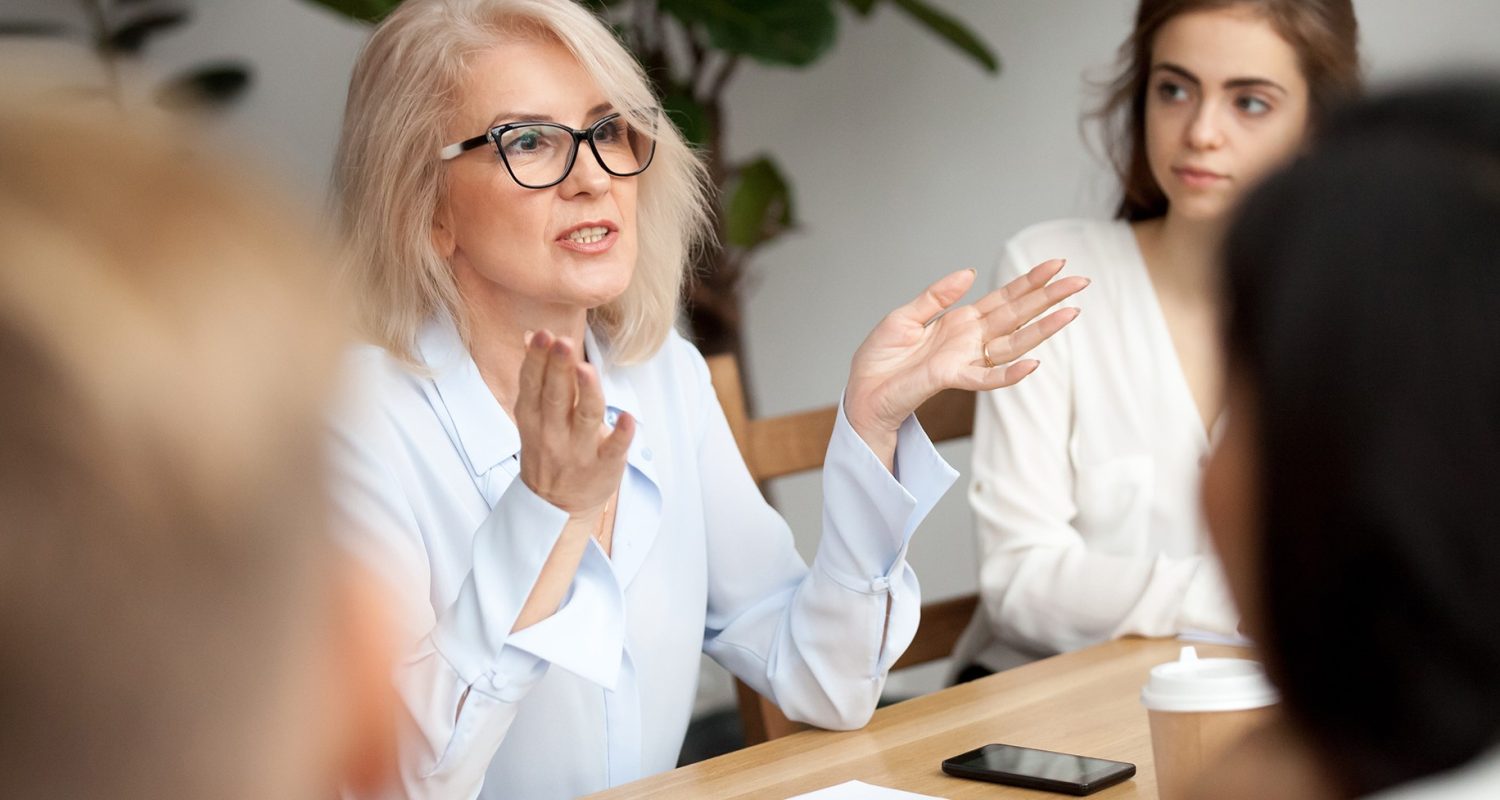 Attractive aged businesswoman, teacher or mentor coach speaking to young people, senior woman in glasses teaching audience at training seminar, female business leader speaker talking at meeting