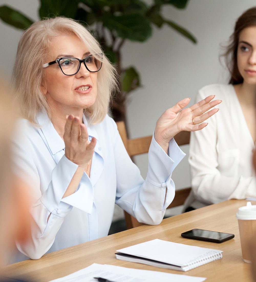 Attractive aged businesswoman, teacher or mentor coach speaking to young people, senior woman in glasses teaching audience at training seminar, female business leader speaker talking at meeting