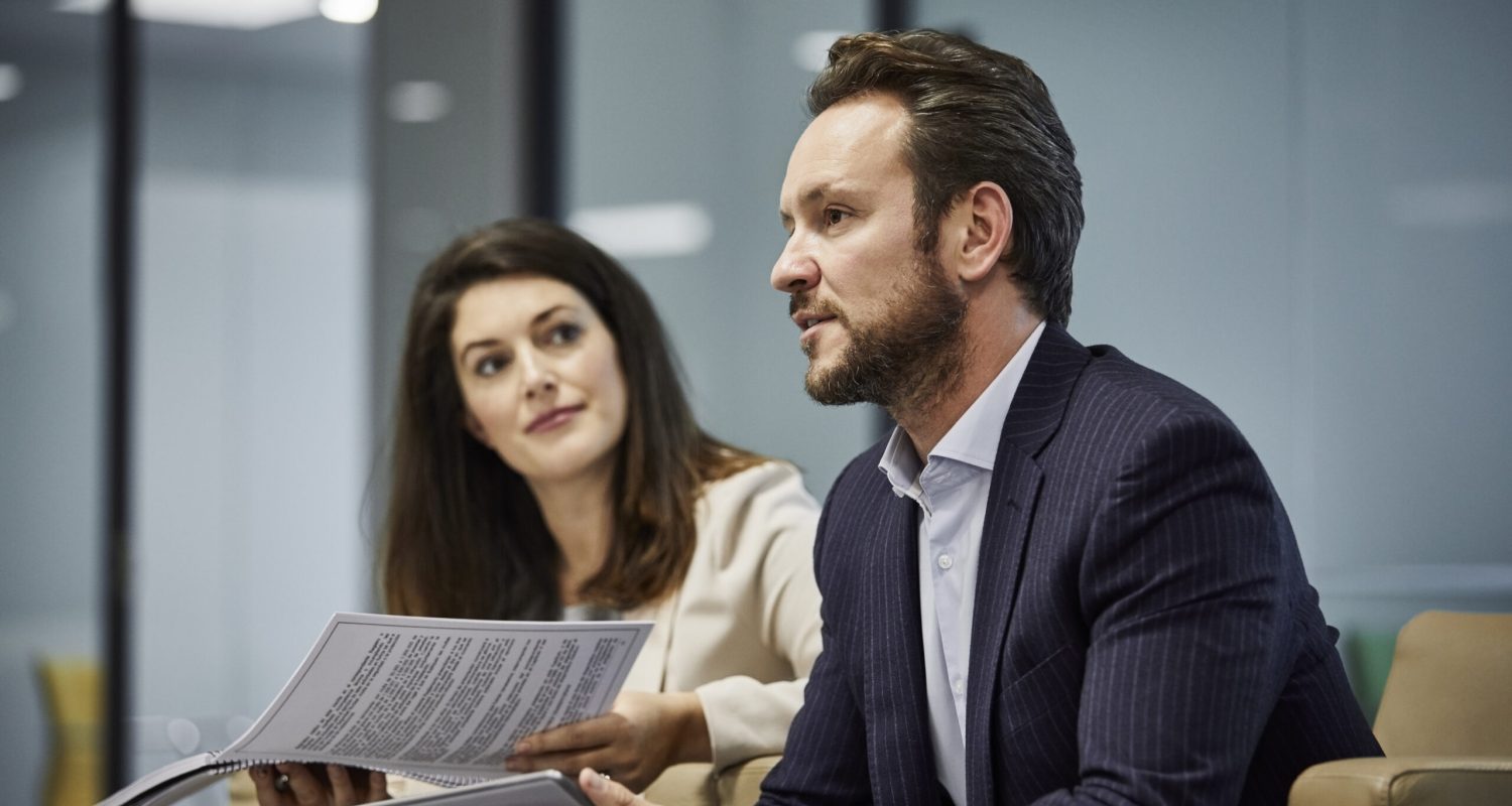 Confident mature businessman holding digital tablet by colleague. Businesswoman is looking at coworker. They are sitting in office lobby.